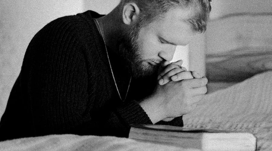 A devoted man in earnest prayer, kneeling beside an open Bible, symbolizing the deep connection between the serenity prayer and the transformative power of faith in addiction recovery.