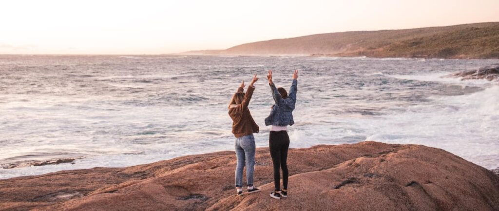 2. Two women celebrating freedom and recovery on a beach, embodying the strength of addiction and relationships healing, with arms joyfully raised to the sky.