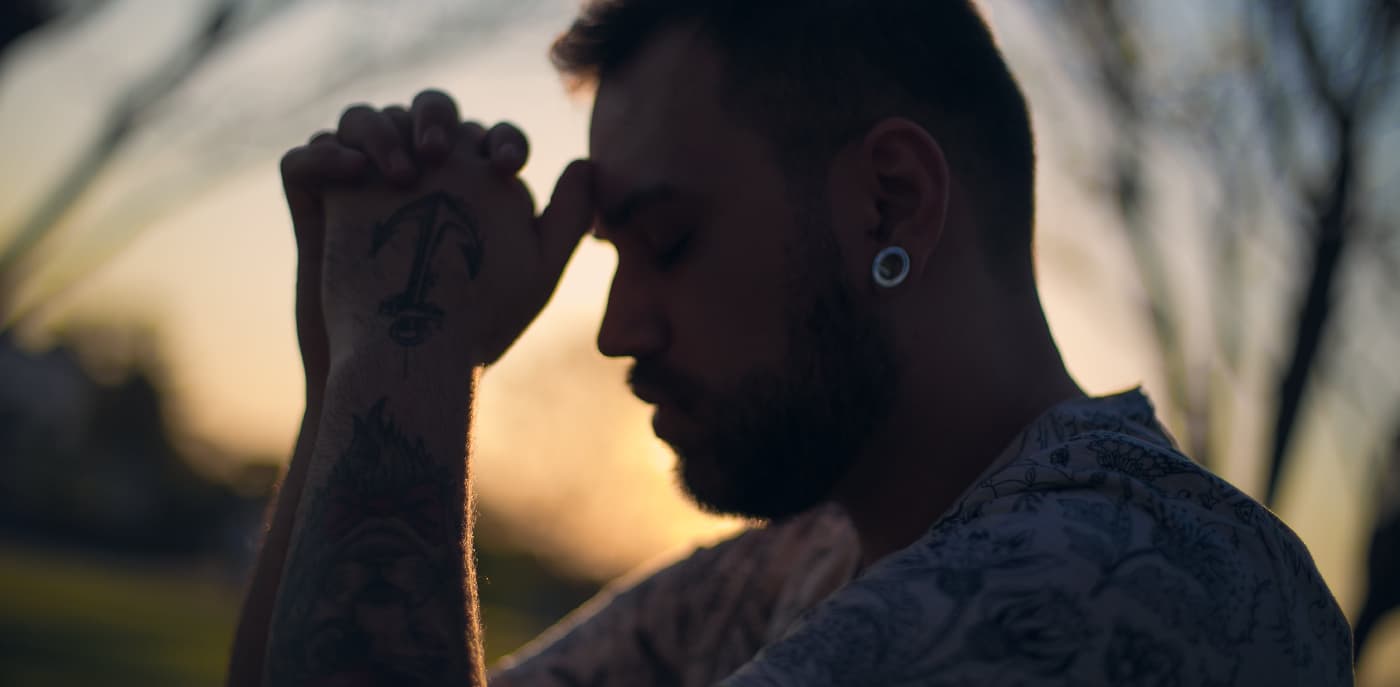 A man with visible tattoos is captured in a moment of prayer, his head bowed and eyes closed, with a serene sunset in the background, symbolizing a journey of humility and recovery.