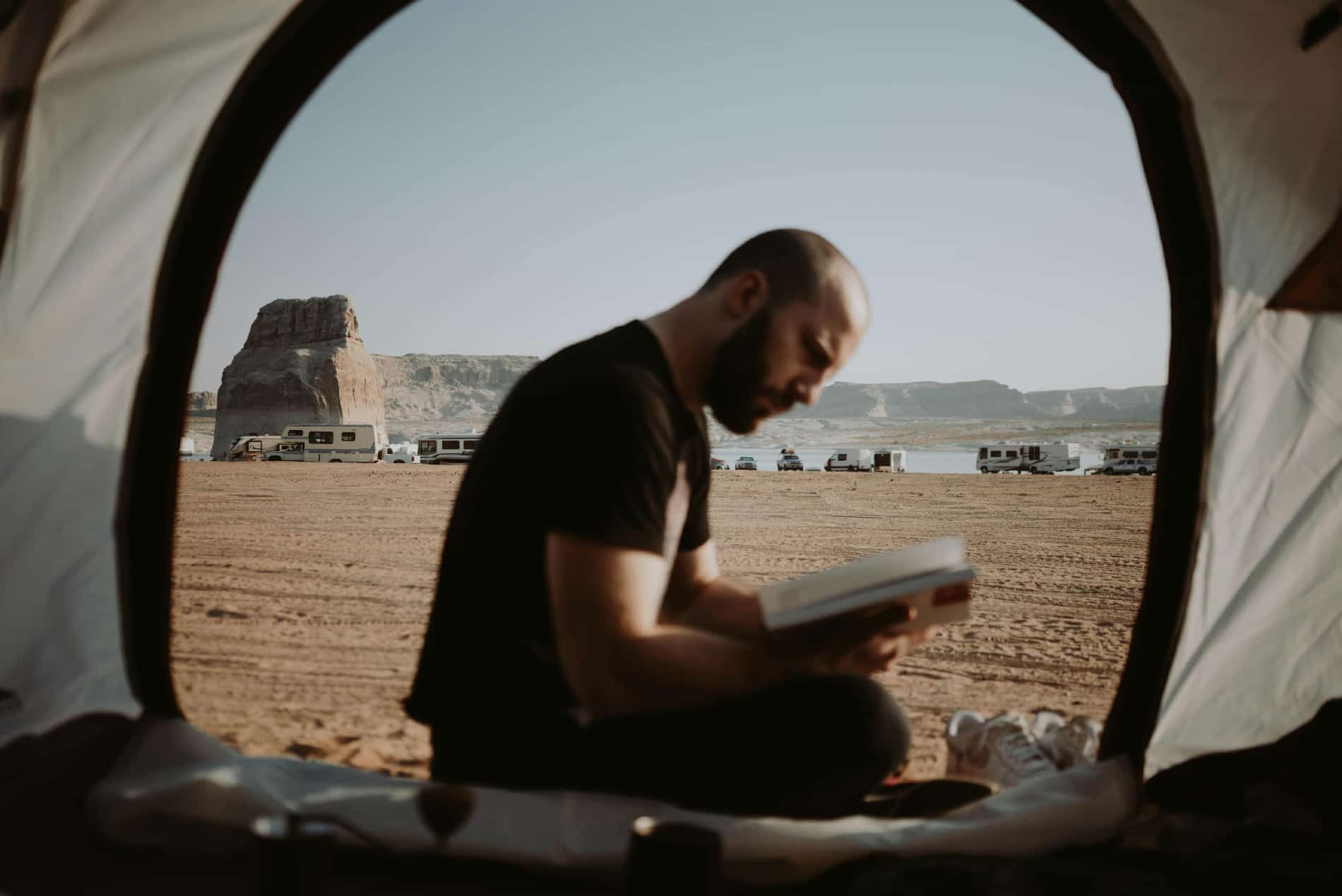 A man sitting beside a tent, absorbed in reading a book with the support of Adderall XR & IR for managing narcolepsy.