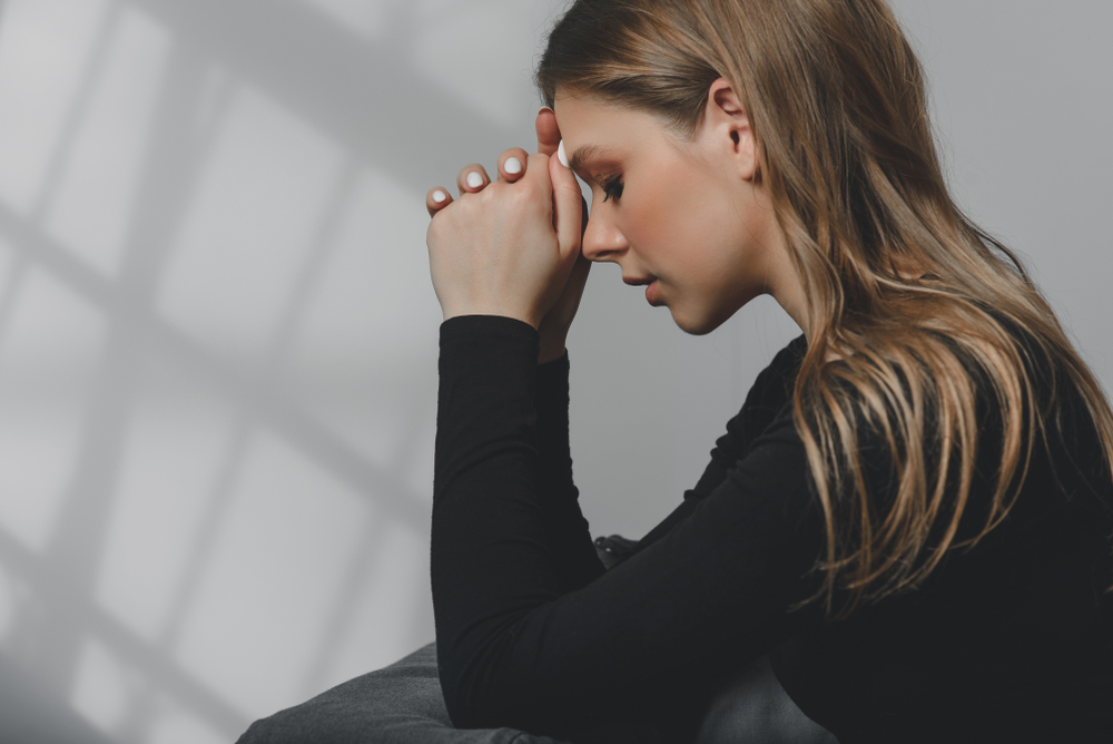 Blonde woman solemnly praying with a gray wall behind her 