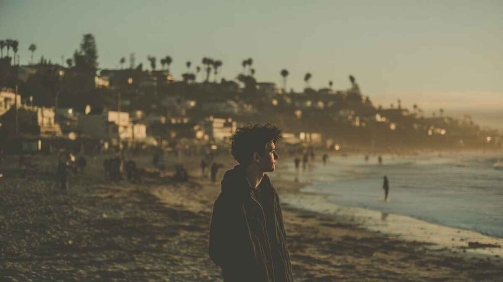 A young man stands alone on a crowded beach at dusk in San Clemente, gazing out at the ocean as warm light softens the shoreline—capturing a quiet moment of contemplation during rehab by the beach.