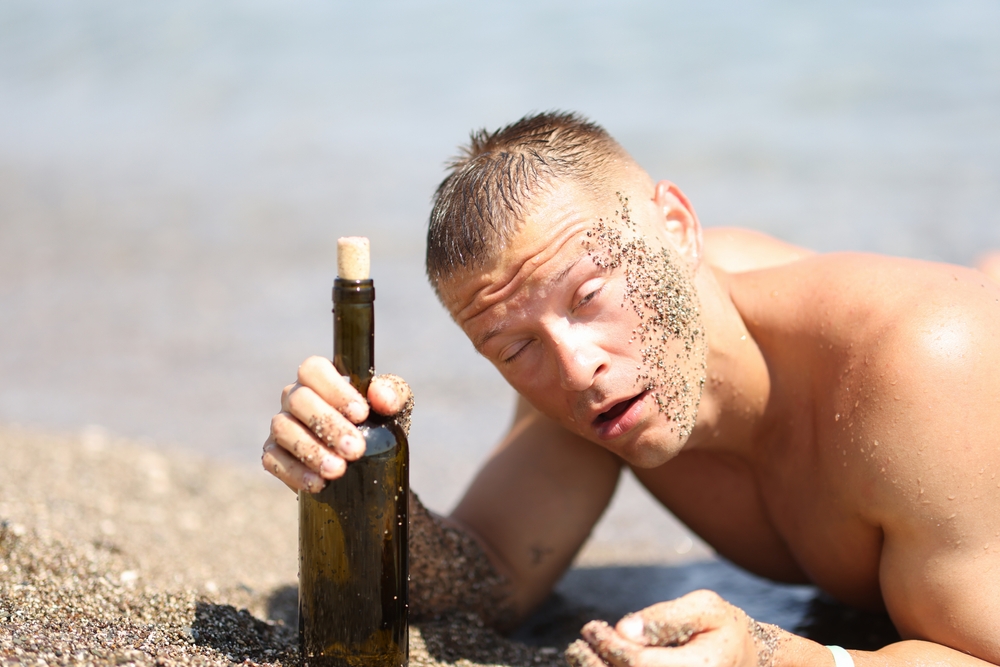 A drunk man on a beach holding an alcohol bottle with eyes sensitive to light